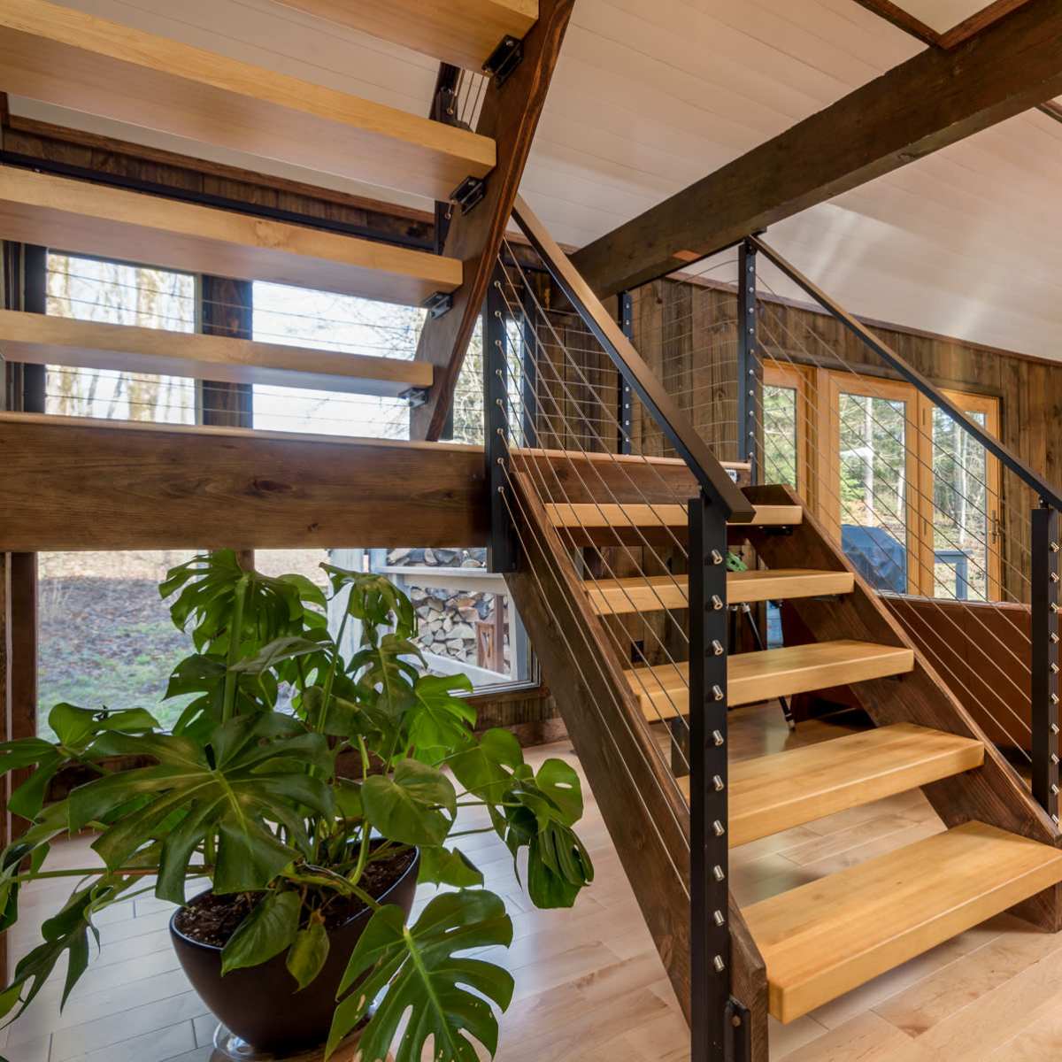 A modern interior with a wooden staircase, a stone fireplace, and large windows overlooking a wooded area.
