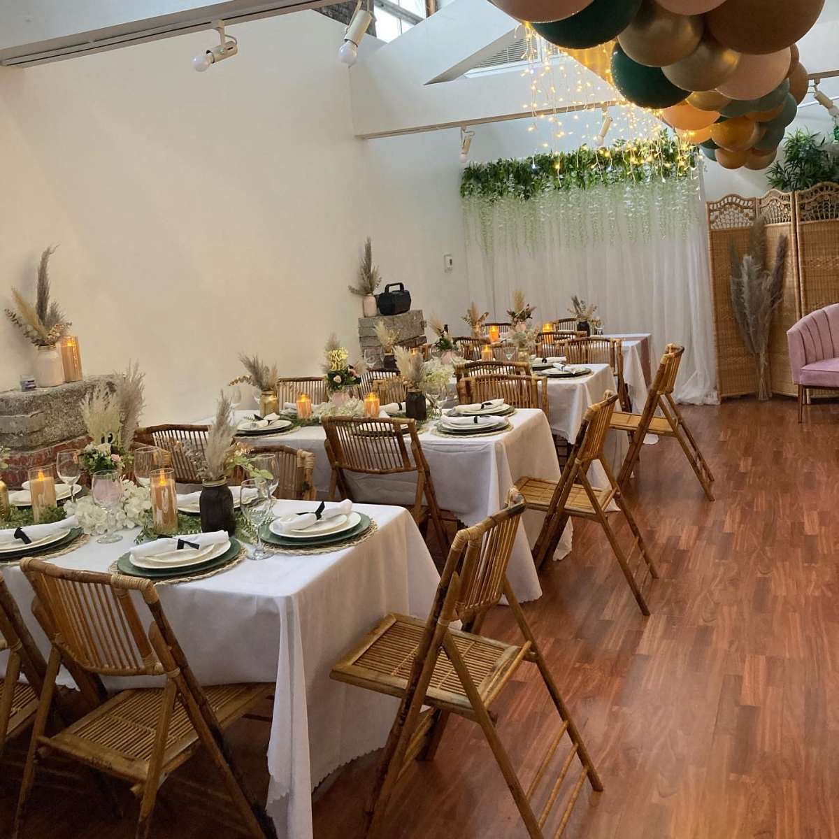 A decorated event space with several tables set for dining, featuring floral centerpieces, candles, and a balloon arrangement hanging above.