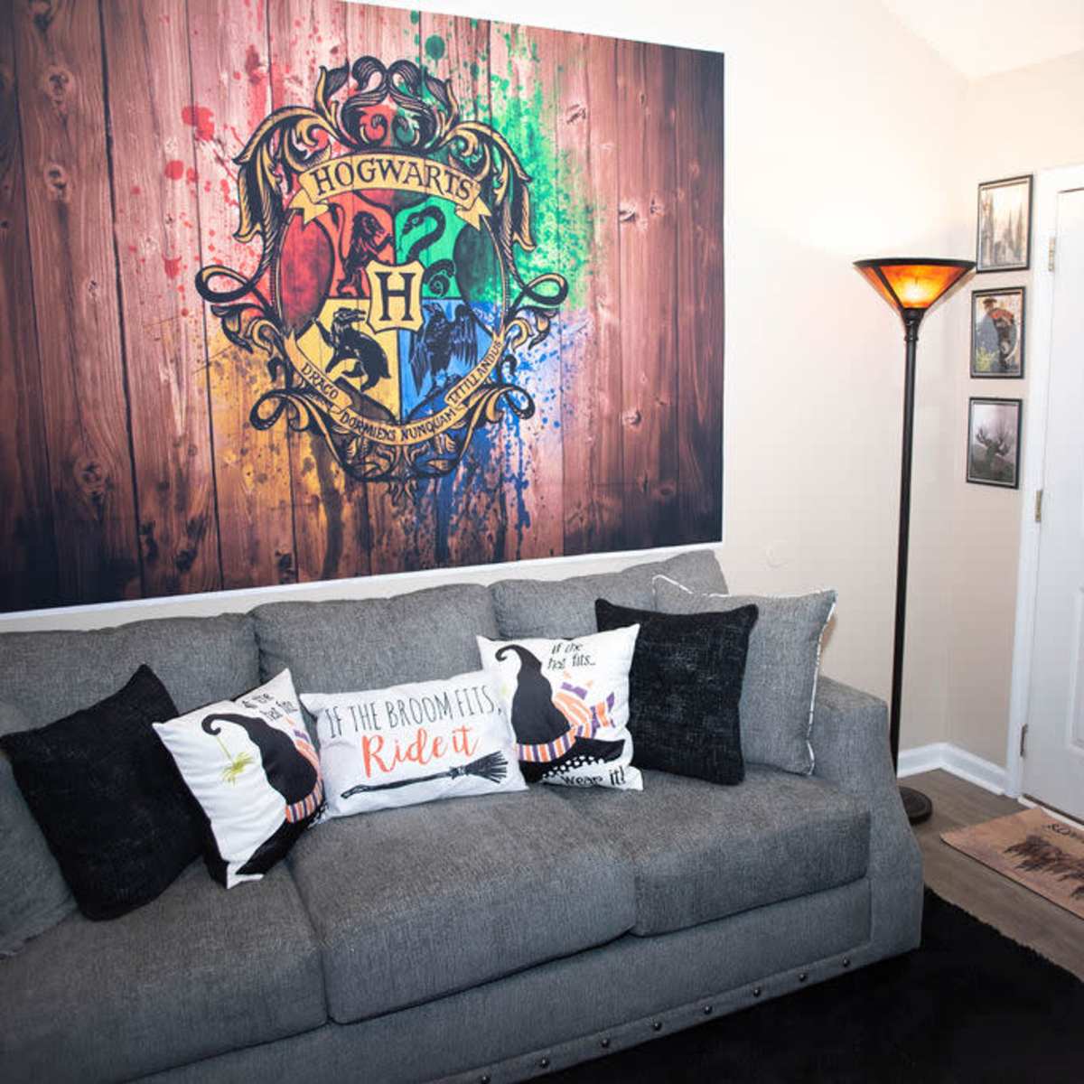 A cozy living room featuring a grey couch with decorative pillows, a large Hogwarts crest mural on the wall, and a front door with a lamp and framed photos nearby.