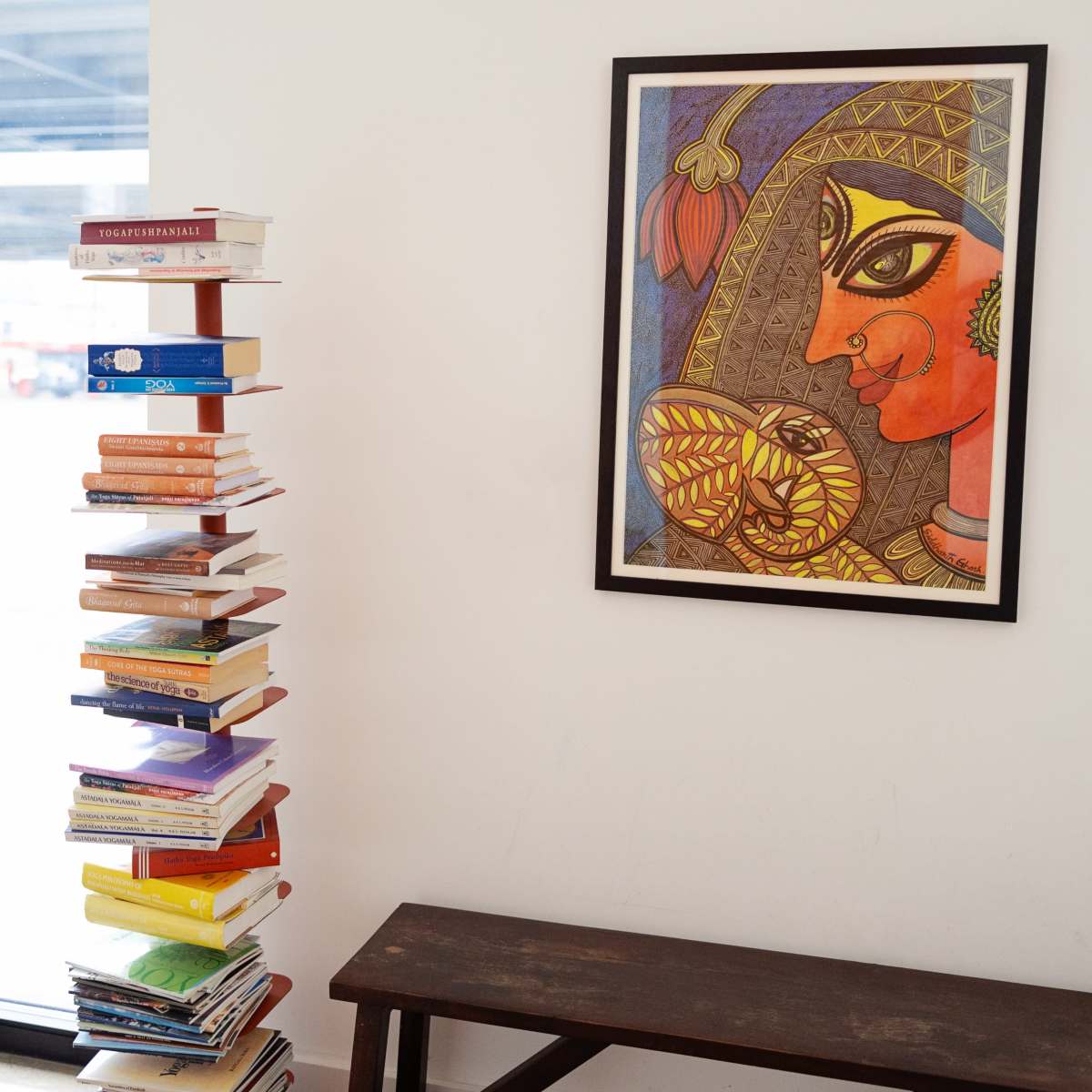 A tall stack of books stands next to a framed painting of a woman with decorative features on a wall.