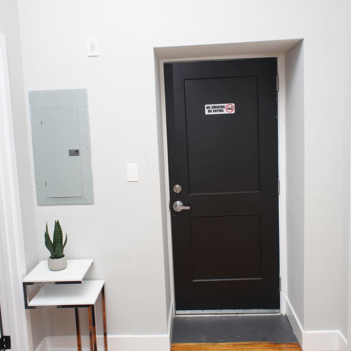 A dark door with a warning sign is located at the end of a hallway, adjacent to a small table with a plant and an electrical panel on the wall.