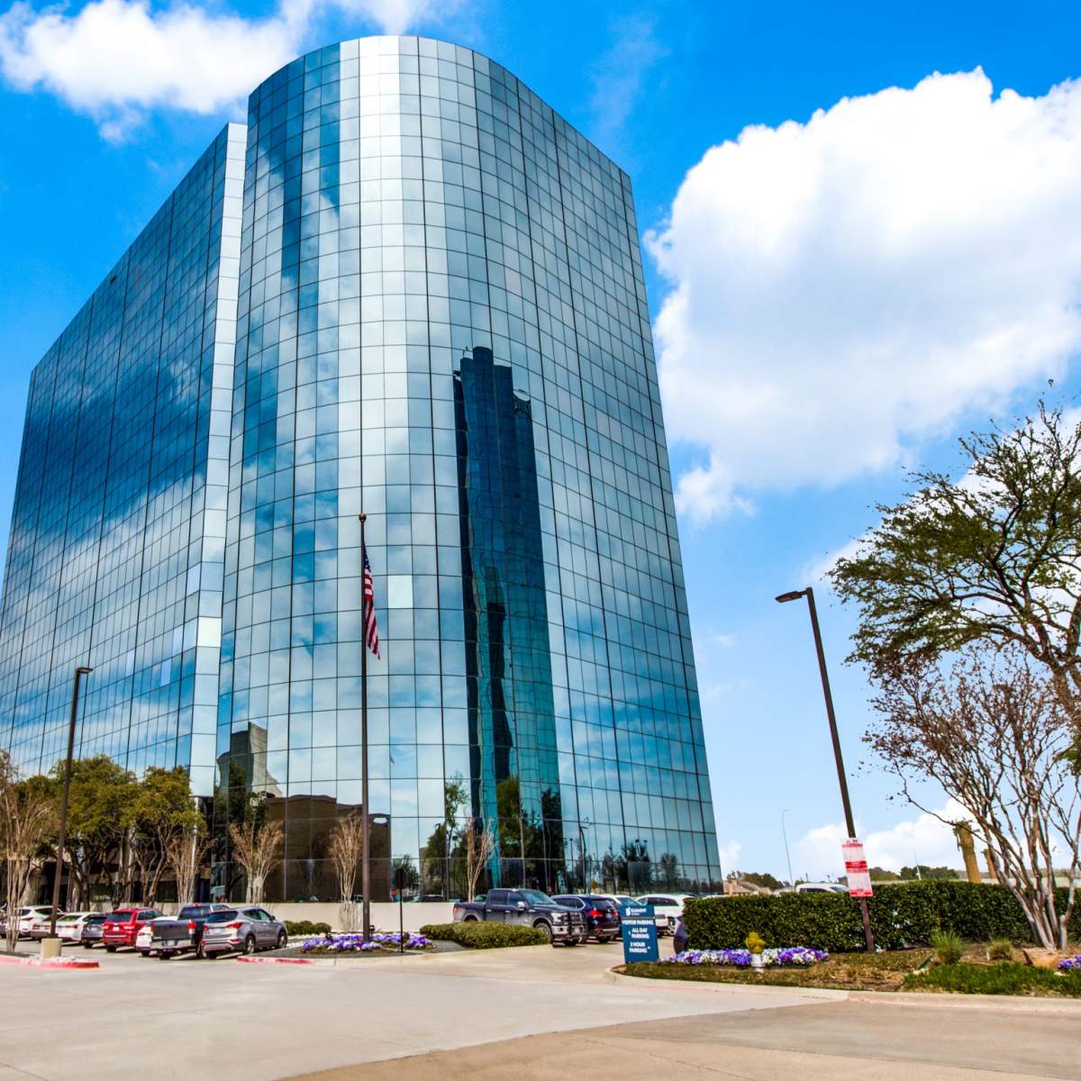 A modern glass building reflects the sky and surrounding trees while featuring an American flag in front.
