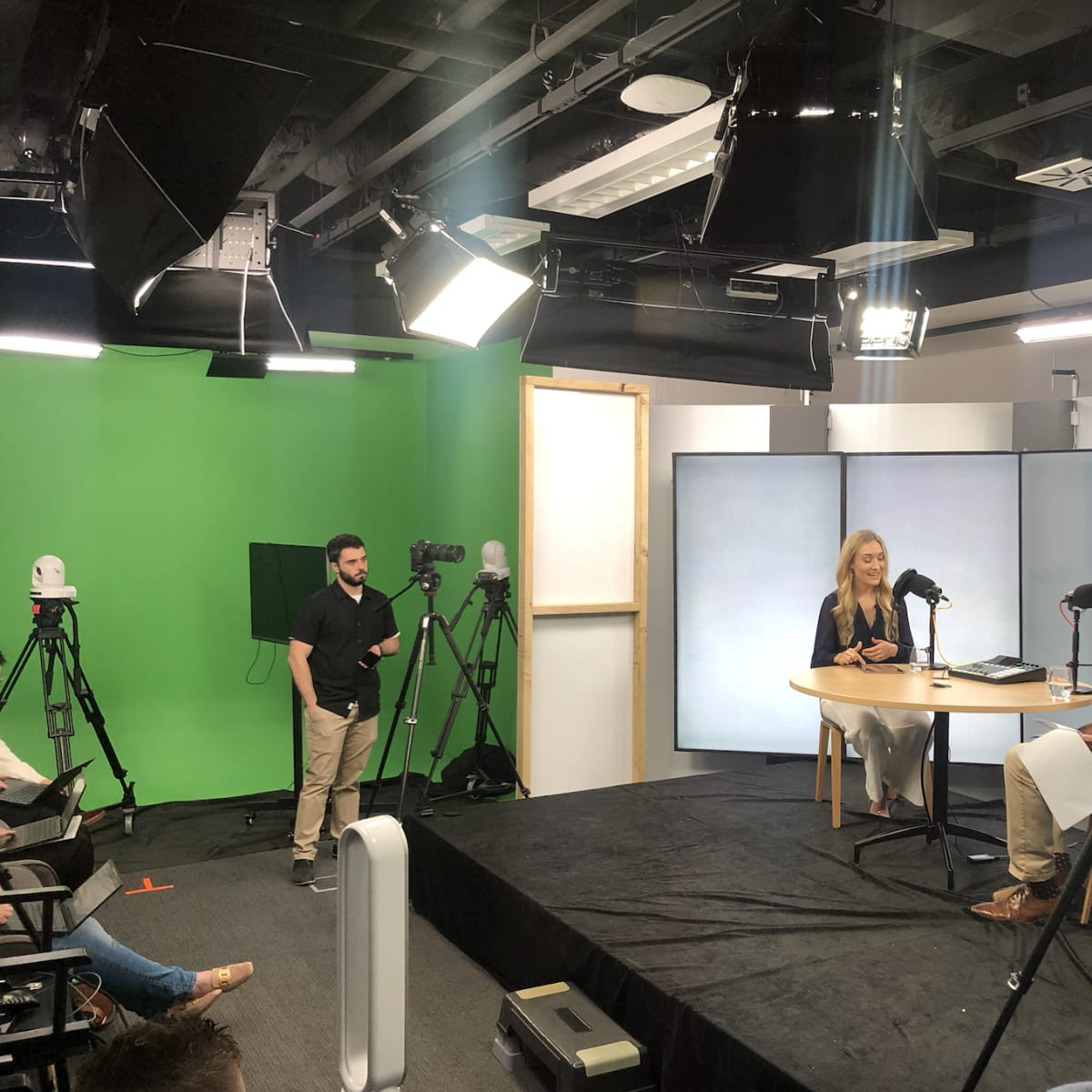 A television studio setting features two people engaged in an interview, with multiple cameras, lighting equipment, and a green screen in the background.