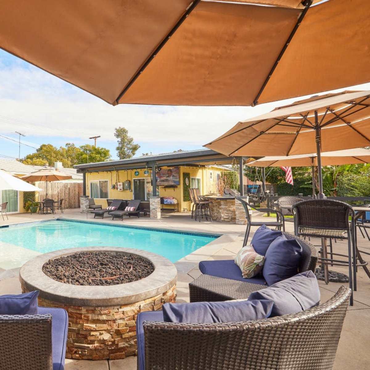 A backyard pool area with a fire pit surrounded by comfortable seating and several umbrellas for shade.