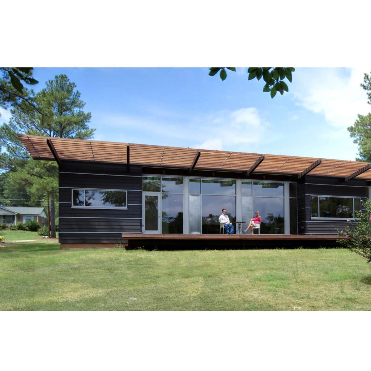 A modern house with large windows and a wooden deck is situated in a grassy area with two people seated outside, surrounded by trees.