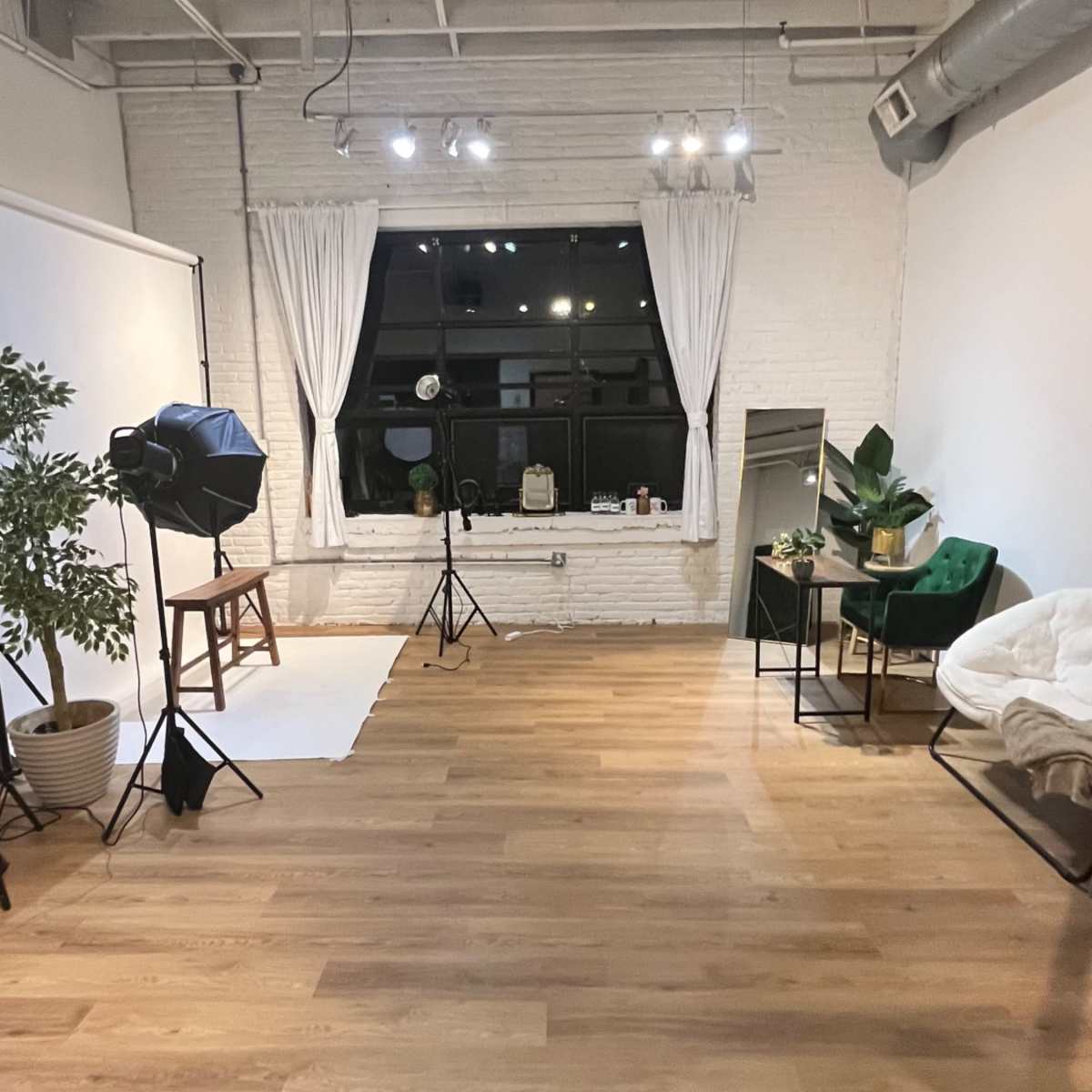 A spacious, well-lit room with wooden flooring, a seating area with green chairs, a backdrop setup for photography, and large windows dressed with white curtains.