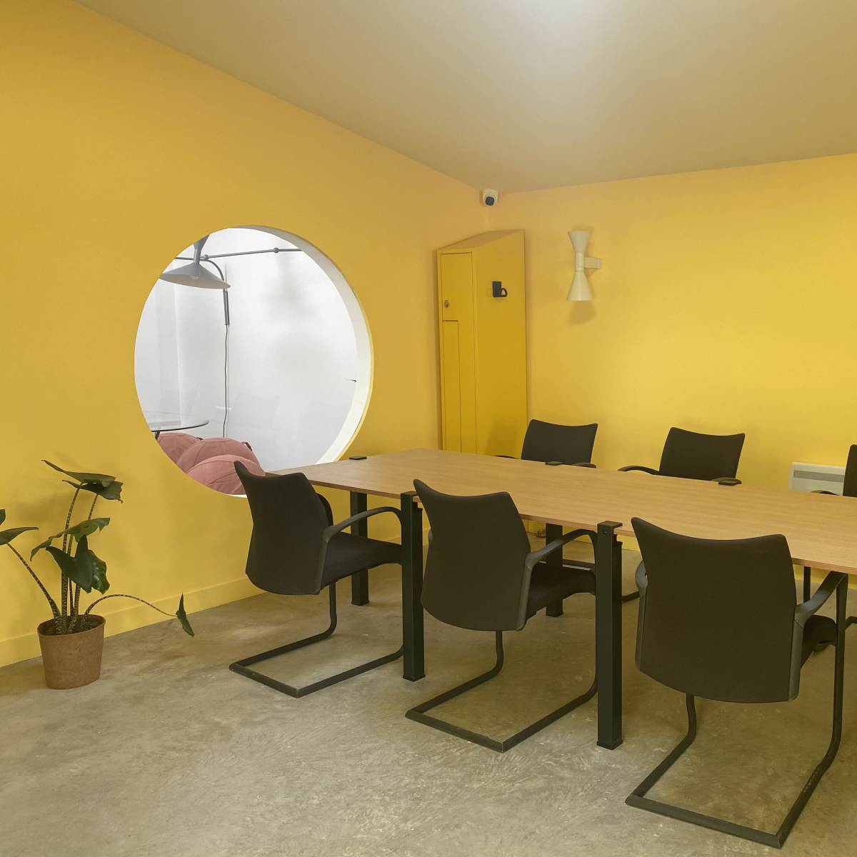 A meeting room with a large yellow wall, a wooden table surrounded by six black chairs, and a circular window leading to another room.