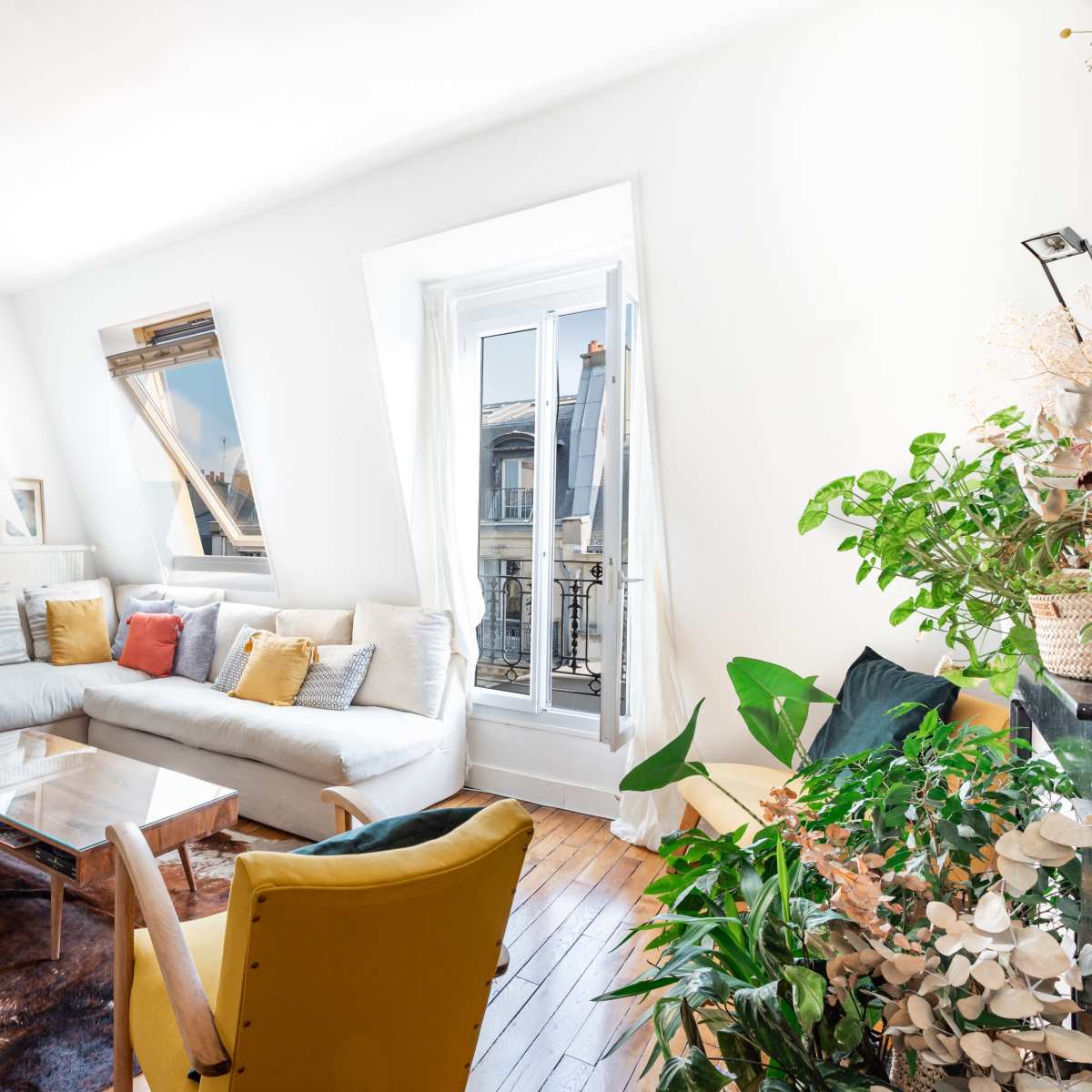 A bright living room features a white sofa, a glass coffee table, and various plants, with a large window that opens to a view of the street.