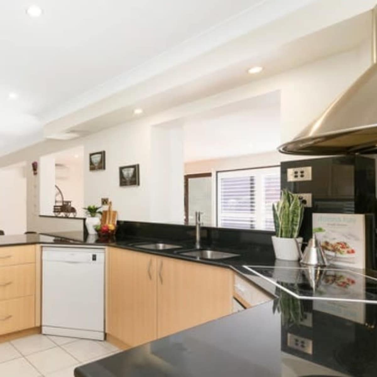 A modern kitchen features light wood cabinetry, black countertops, and stainless steel appliances, with a view into a living area in the background.
