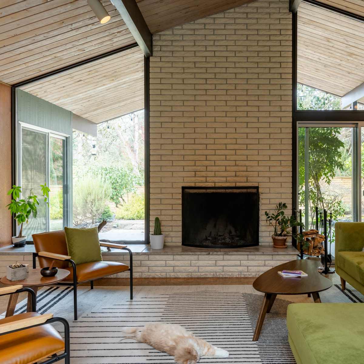 A modern living room features a stone fireplace, large windows with views of greenery, and several seating options including a green sofa and brown chairs.