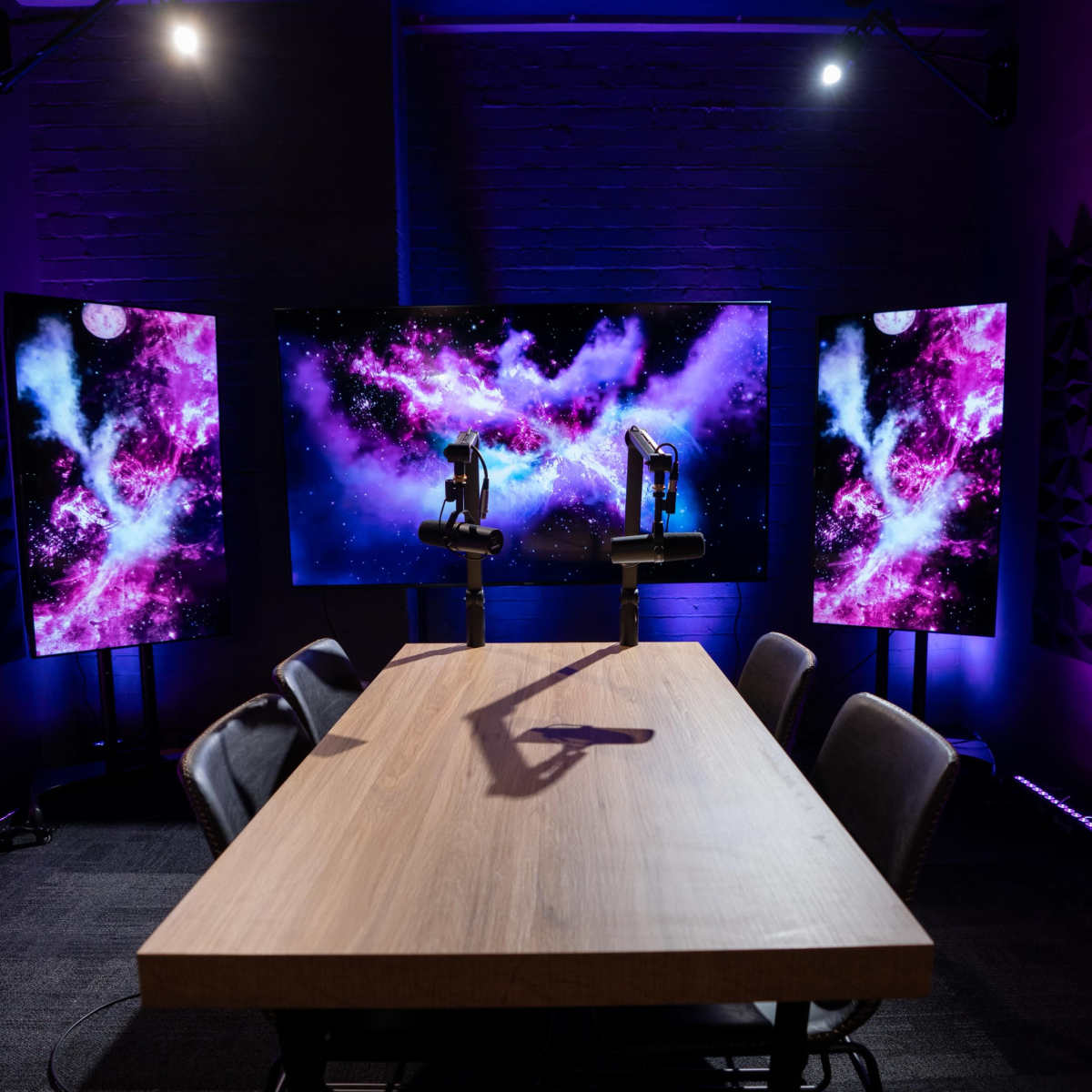 A conference room features a wooden table with black chairs, flanked by two large screens displaying a colorful cosmic background.