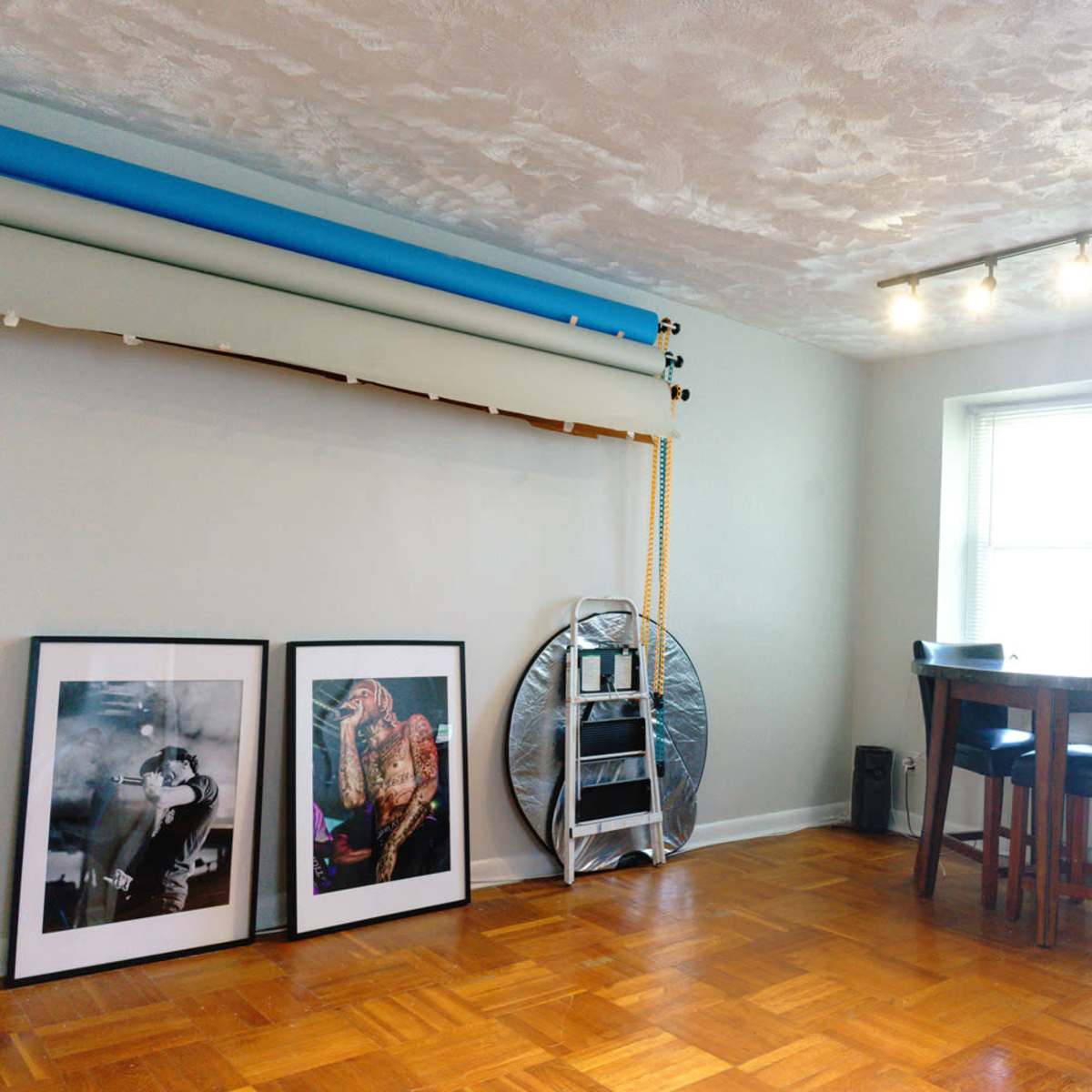 A minimalistic room featuring two framed photographs on the floor, a backdrop setup, a round table with chairs, and a window with multiple light sources.