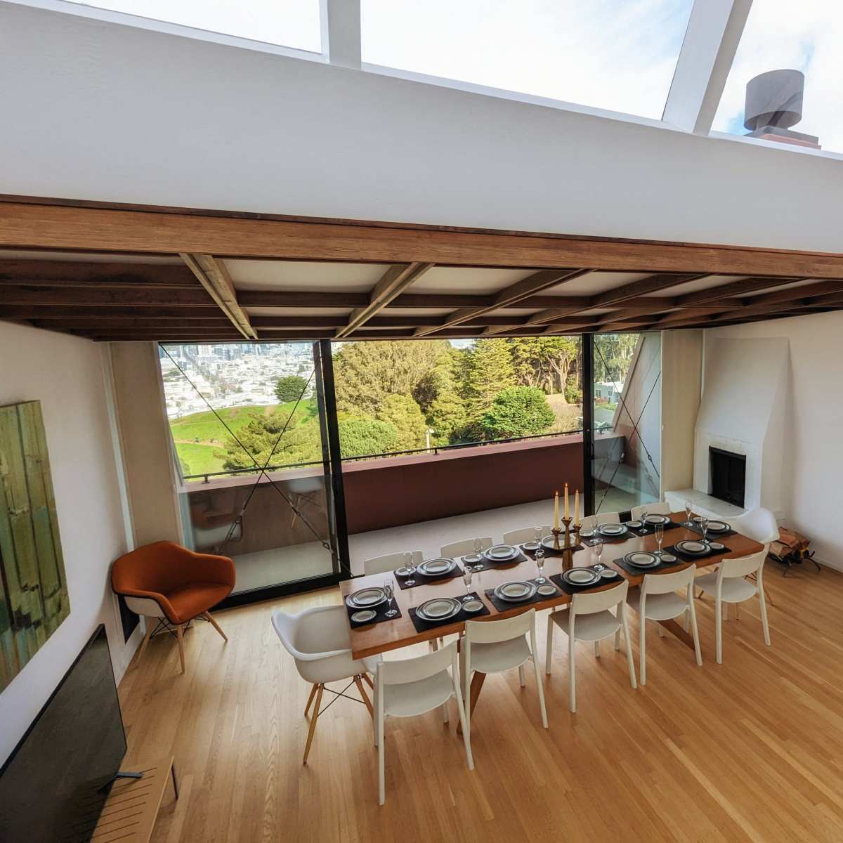 A dining room with a long wooden table set for a meal, surrounded by white chairs and large windows that provide a view of lush greenery outside.