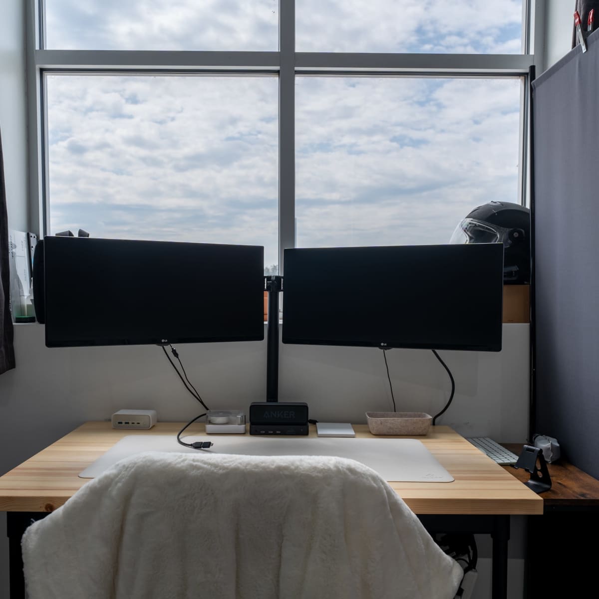 A desktop setup featuring two monitor screens, a light-colored desk with a keyboard and mouse, and a window with a cloudy sky in the background.