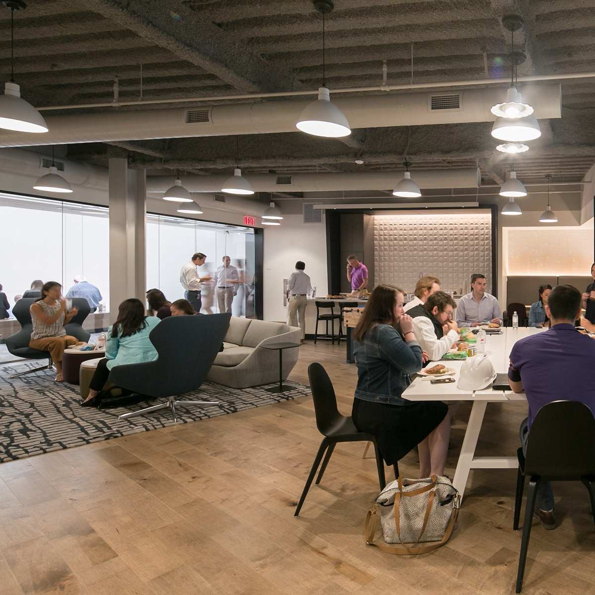 A group of people is gathered in a modern office space, participating in discussions and working at tables while others mingle in the background.