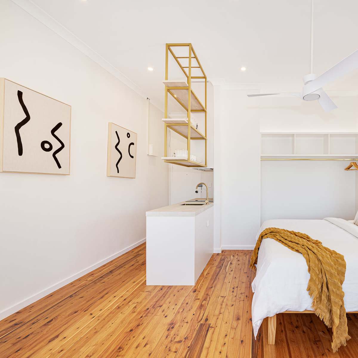A bright, modern bedroom featuring a large bed with white linens, wooden flooring, abstract wall art, and a small kitchenette against one wall.