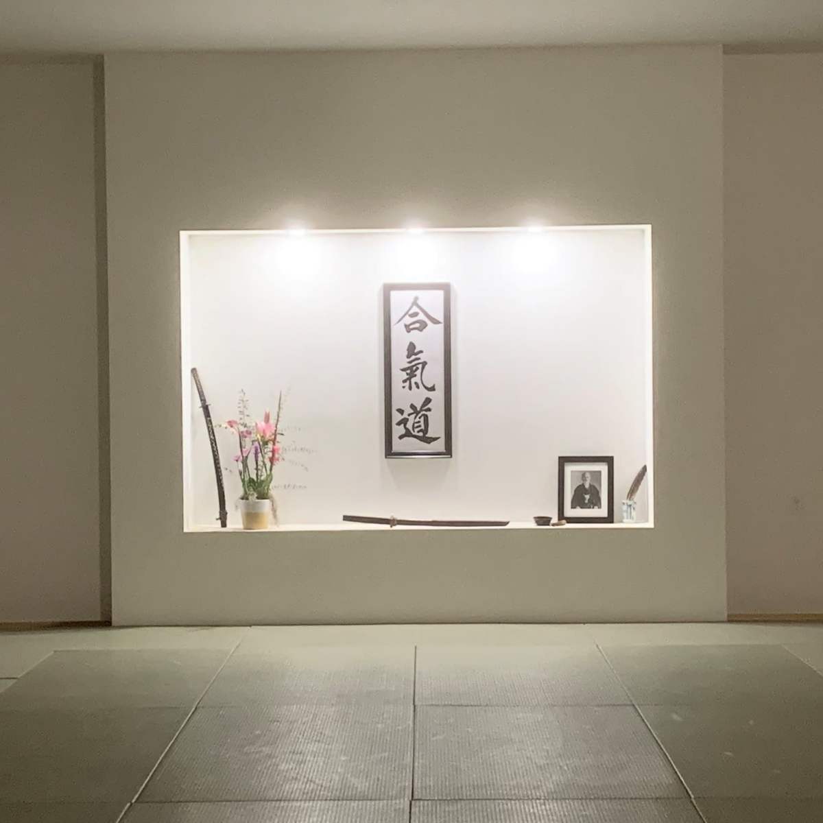 A minimalist display features a wall niche with a calligraphic scroll, a potted plant, a framed photograph, and two traditional Japanese swords.