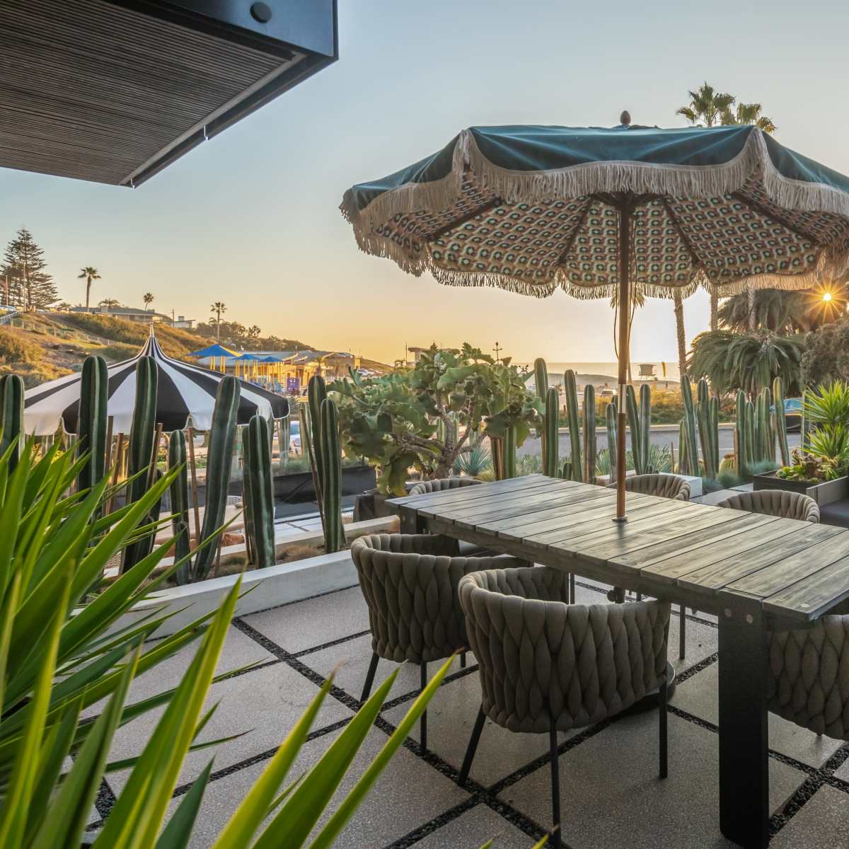 A patio area features a large umbrella over a dining table surrounded by woven chairs, with palm trees and a building in the background illuminated by sunset.