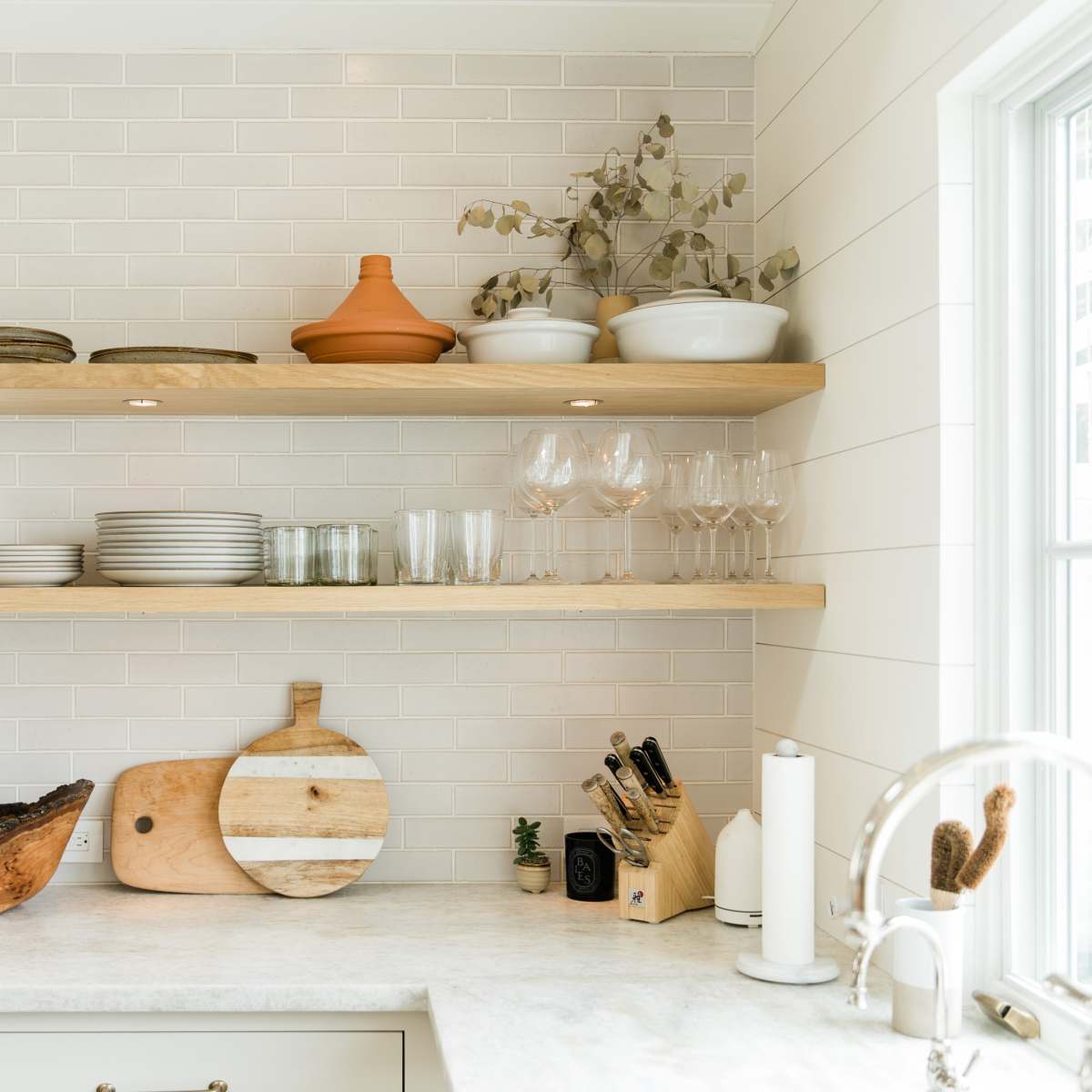 A modern kitchen with open wooden shelves displaying plates, glasses, and various serving bowls, alongside a countertop with cutting boards and kitchen utensils.