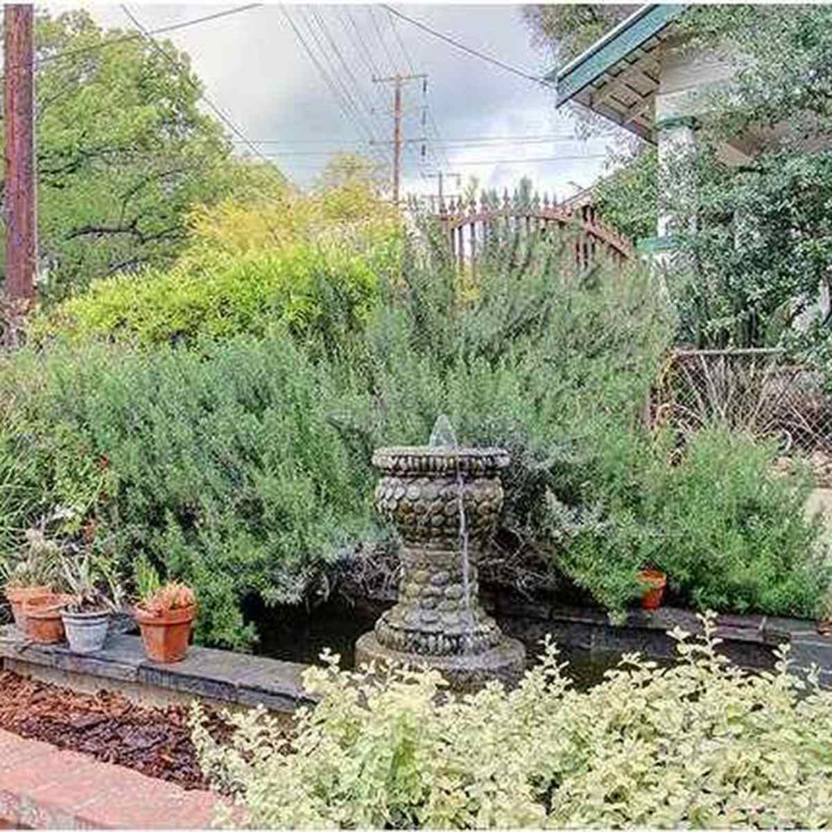 A stone fountain sits amidst lush greenery and potted plants in a garden enclosed by a wrought iron fence.