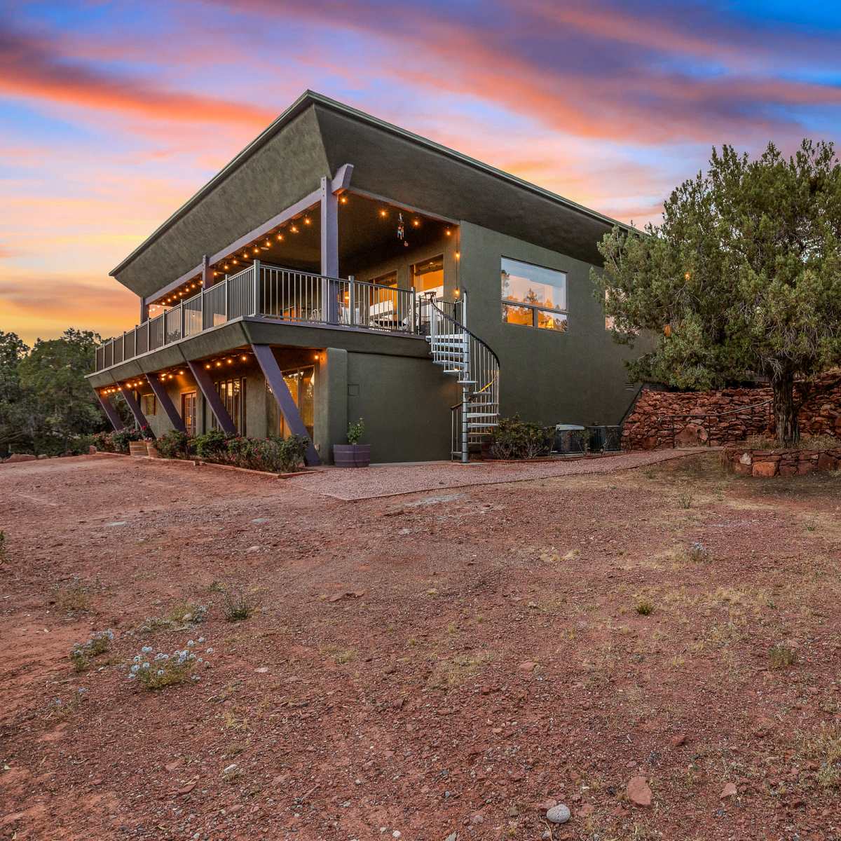 A modern two-story house with a green exterior is situated on a gravel lot, surrounded by trees and rocky landscaping, with a fire pit area nearby.