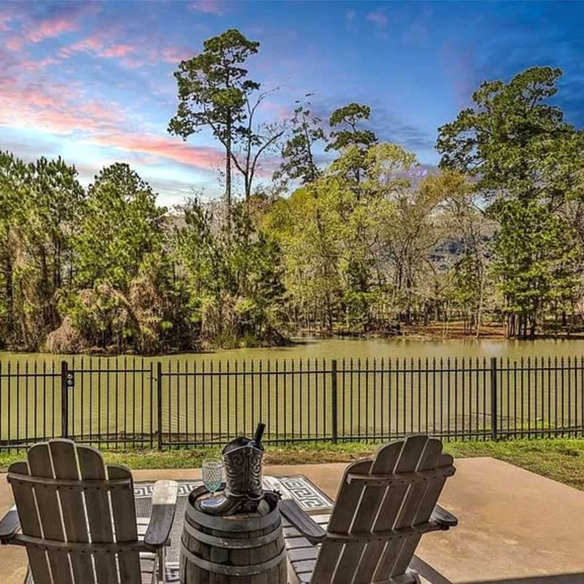 A patio with two chairs and a table overlooks a tranquil body of water surrounded by trees.