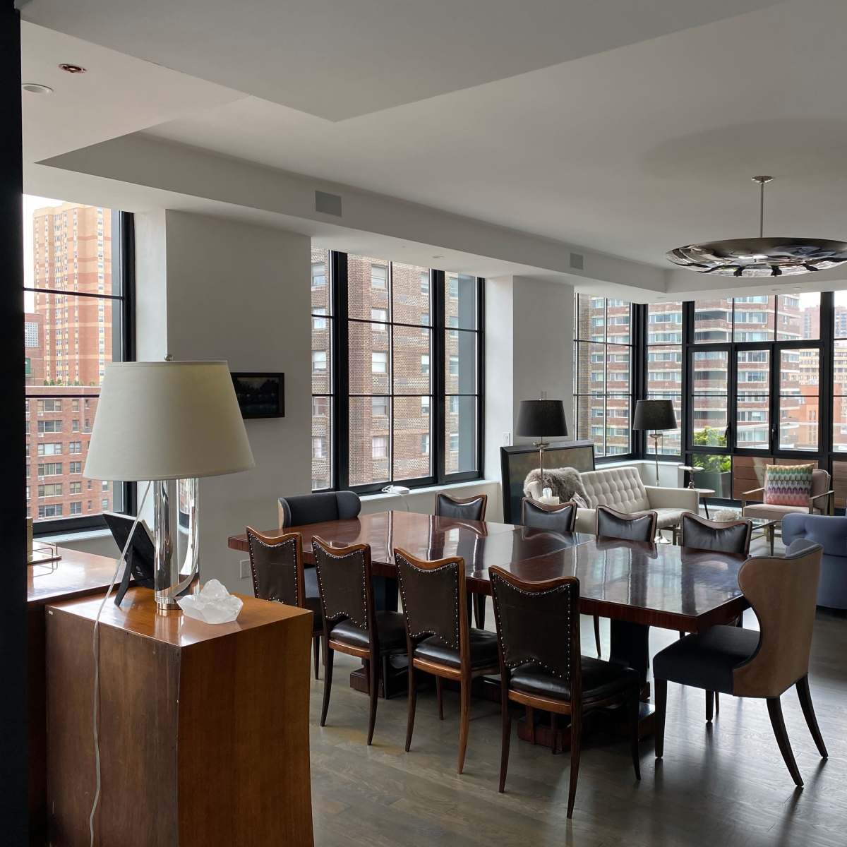 A spacious, modern living area features a large dining table surrounded by chairs, with multiple windows providing views of a city skyline.