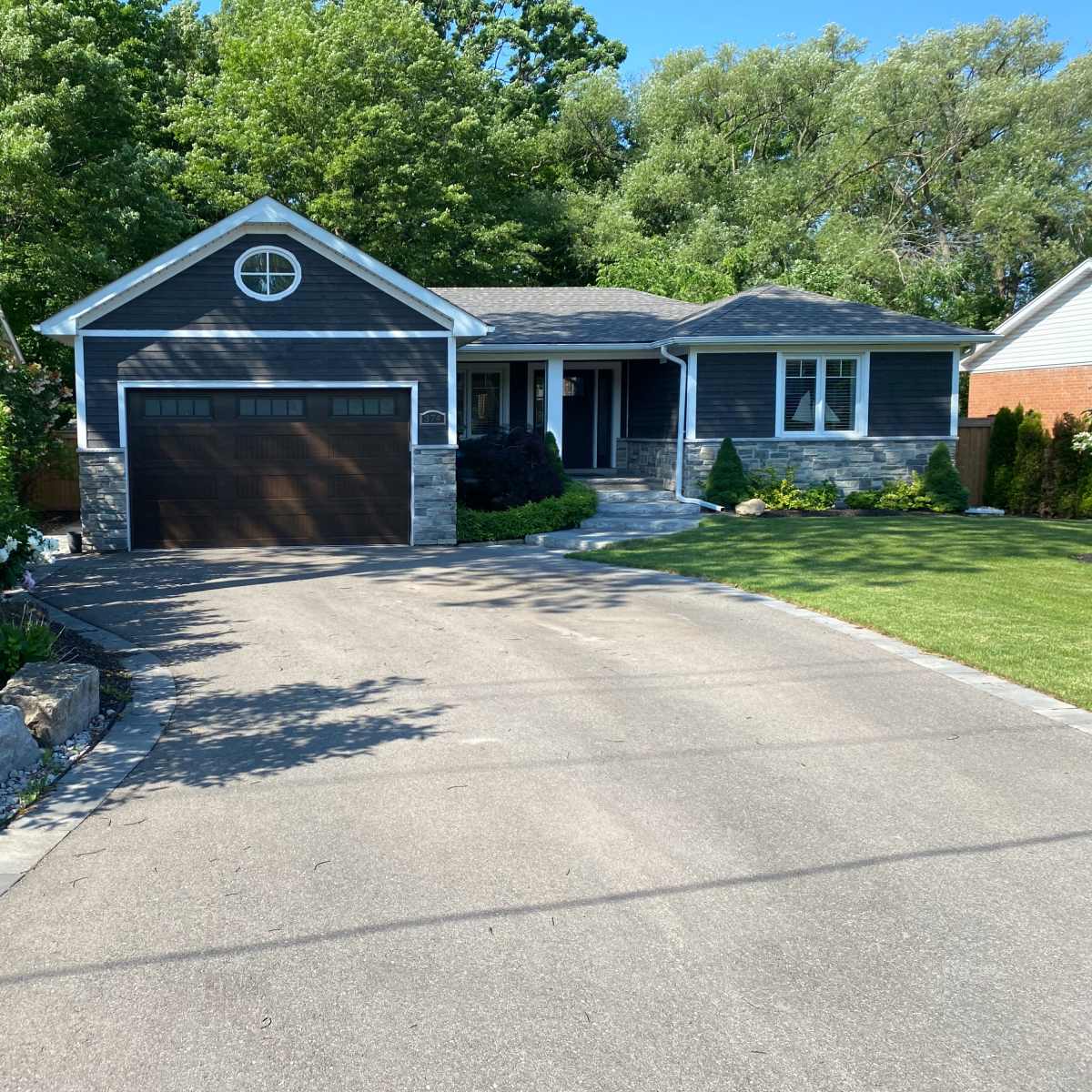 A single-story house with a dark gray exterior and a brown garage door is set along a paved driveway surrounded by greenery.