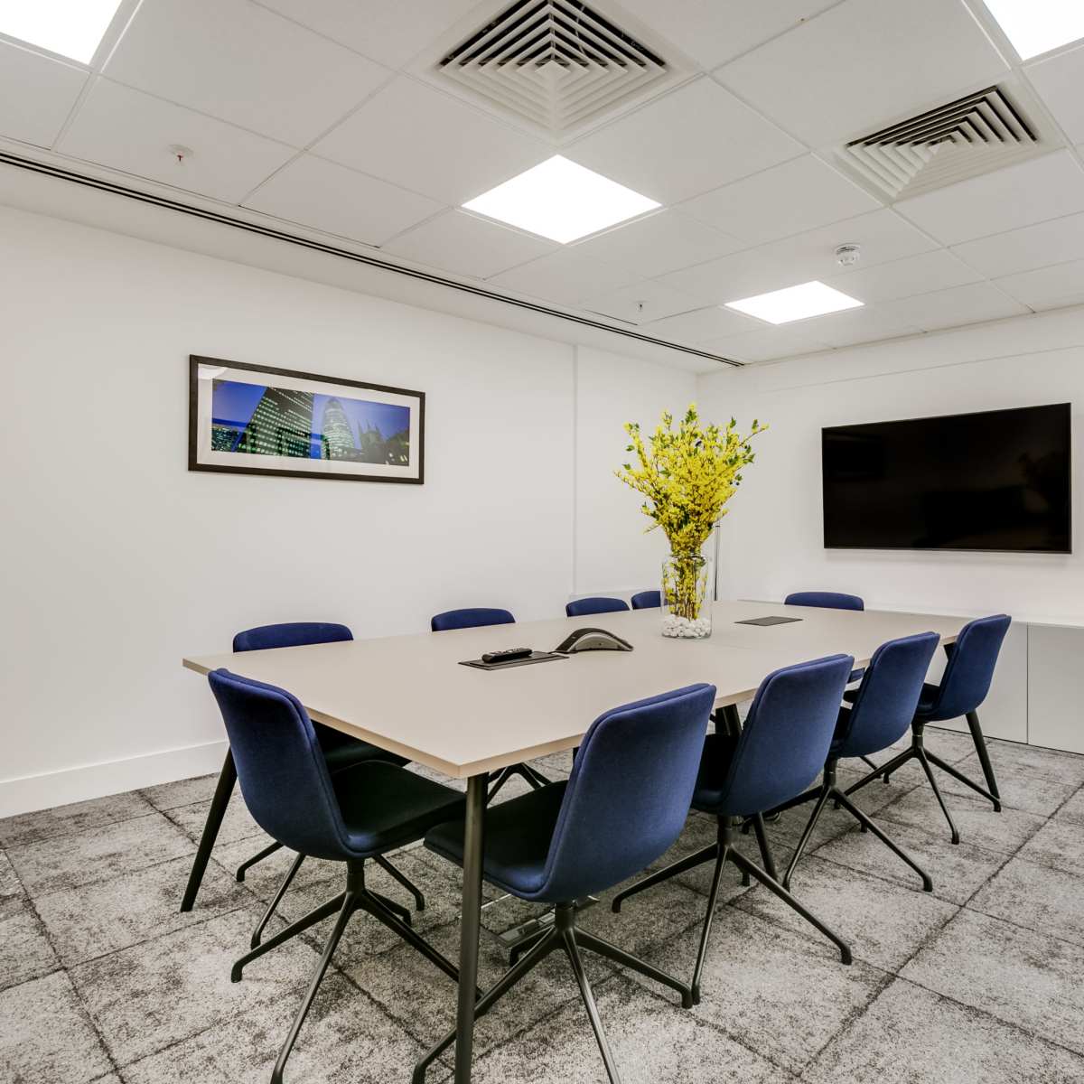 A modern conference room features a large rectangular table surrounded by blue chairs, with a television mounted on the wall and a vase of yellow flowers as a centerpiece.