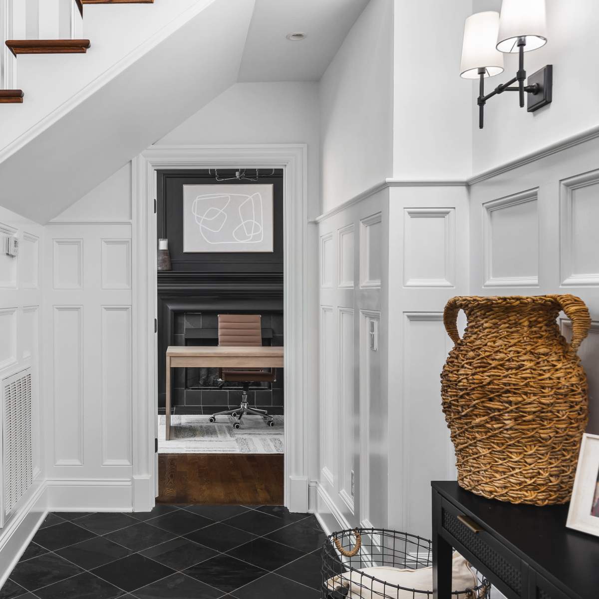 A well-lit hallway featuring a staircase, white paneling, and a view into a room with a black fireplace and modern decor.
