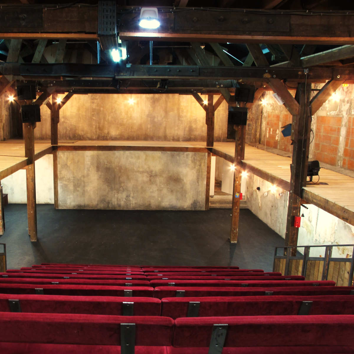 An empty theater with red seating facing a performance stage, featuring exposed wooden beams and a rustic interior.