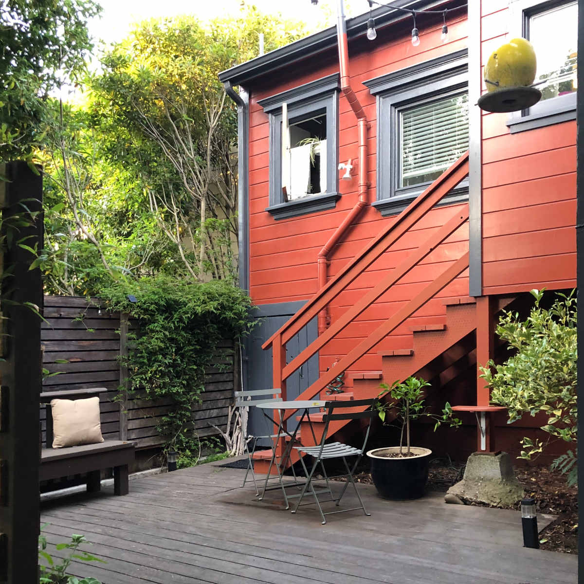 A small outdoor area with a wooden deck, a seating arrangement, and a red house with a staircase and green foliage surrounding it.