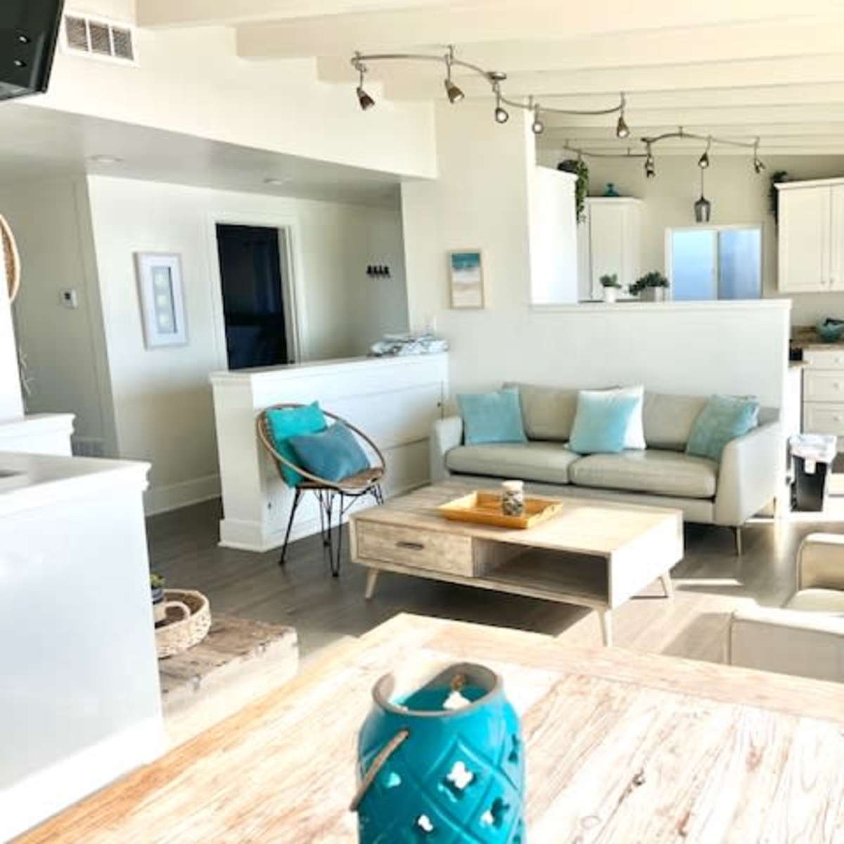 A bright living room with light-colored furniture, a wooden coffee table, and an open kitchen space in the background.