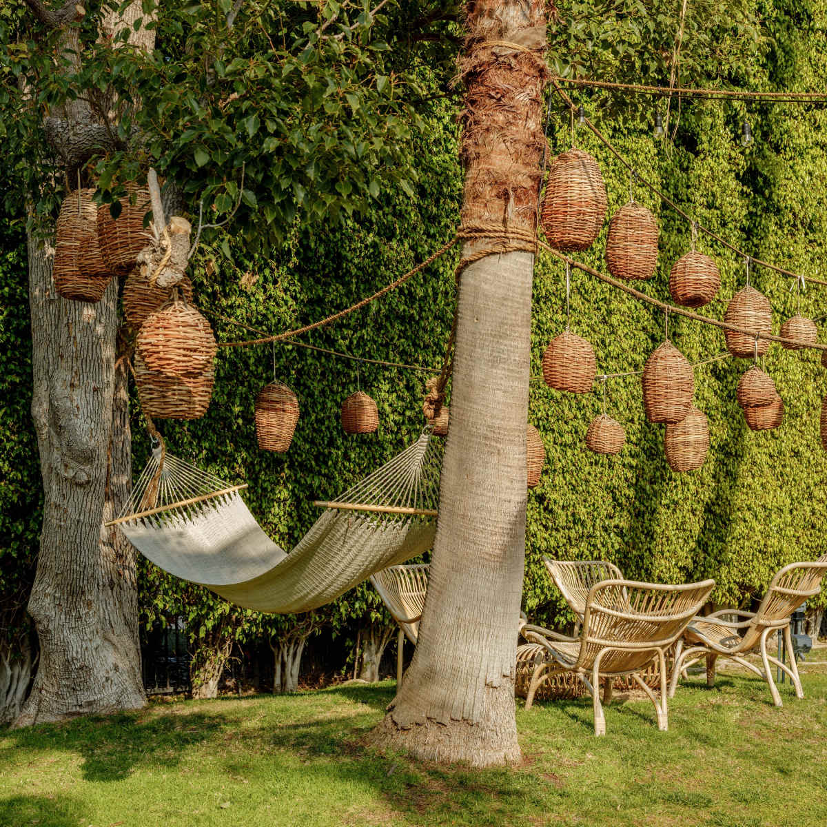 A hammock is suspended between two trees, with woven baskets hanging from branches and lounge chairs arranged on the grass.