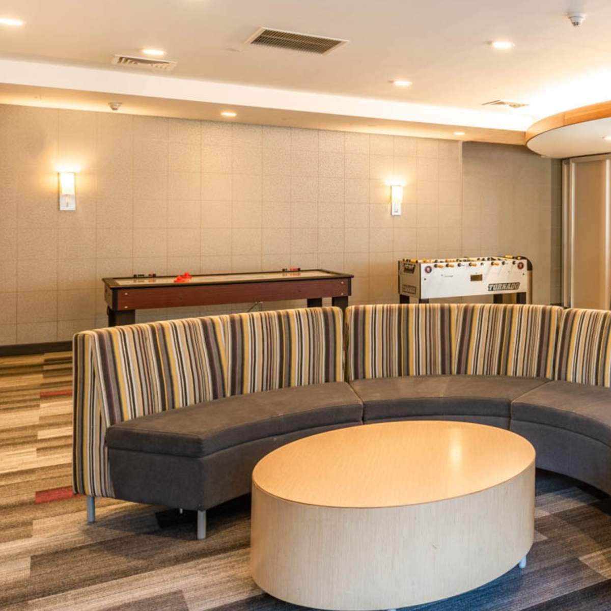 A modern lounge area featuring a curved striped seating arrangement with a circular wooden table, a game table, and a bar counter with stools in a spacious room.