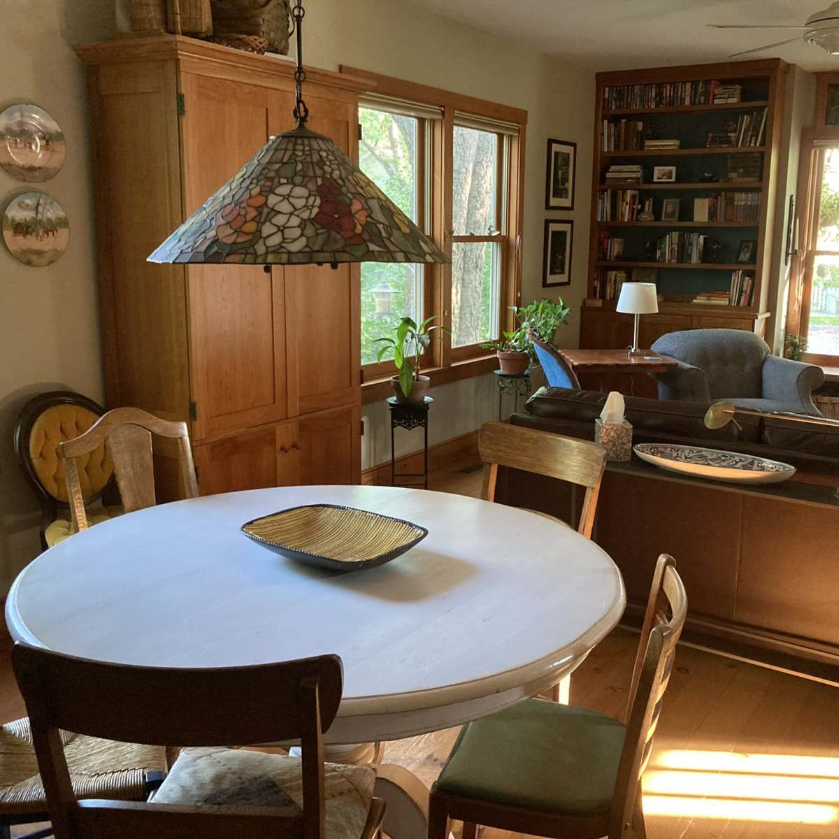 A dining area with a round white table surrounded by chairs, leading into a living room filled with bookshelves and large windows.