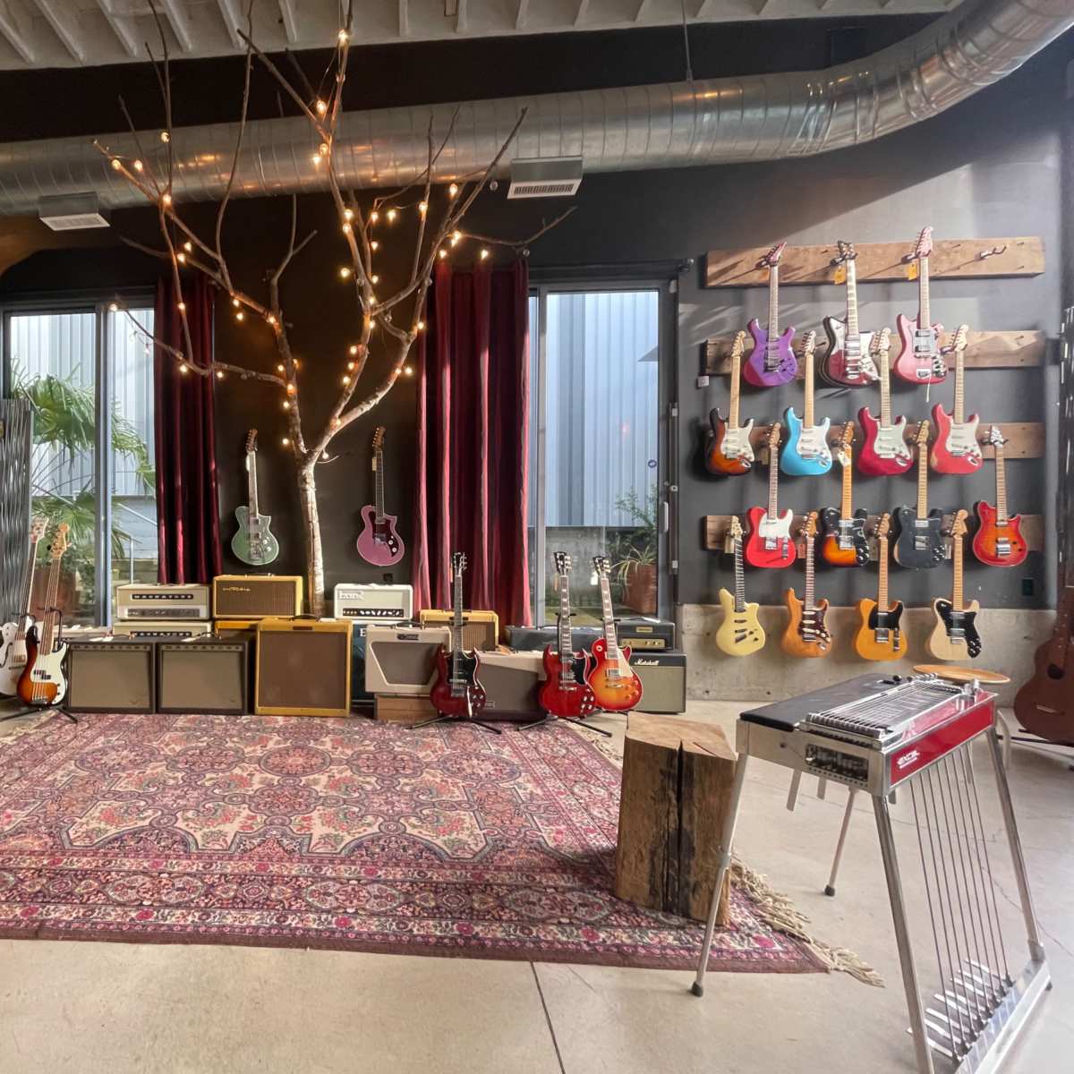 A music studio featuring an array of guitars displayed on walls, amplifiers arranged in a seating area, and a vintage carpet on the floor, complemented by decorative lights and a tree branch.