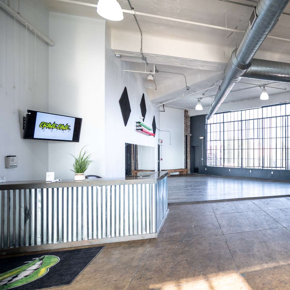 An industrial-style event space featuring a large window, brick walls, a small stage with a microphone, and empty wooden flooring.