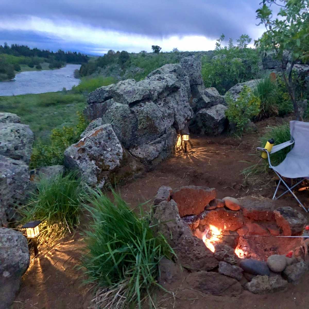 A small, rocky campsite features a fire pit surrounded by two chairs and lanterns, with a river and greenery in the background.