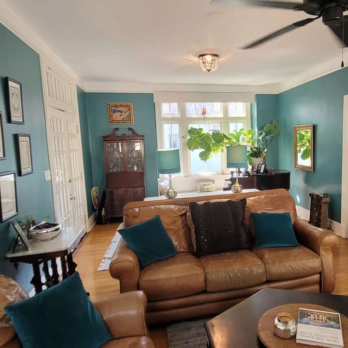 A living room with teal walls, featuring a mix of leather and fabric seating, a coffee table, and a large window allowing natural light.