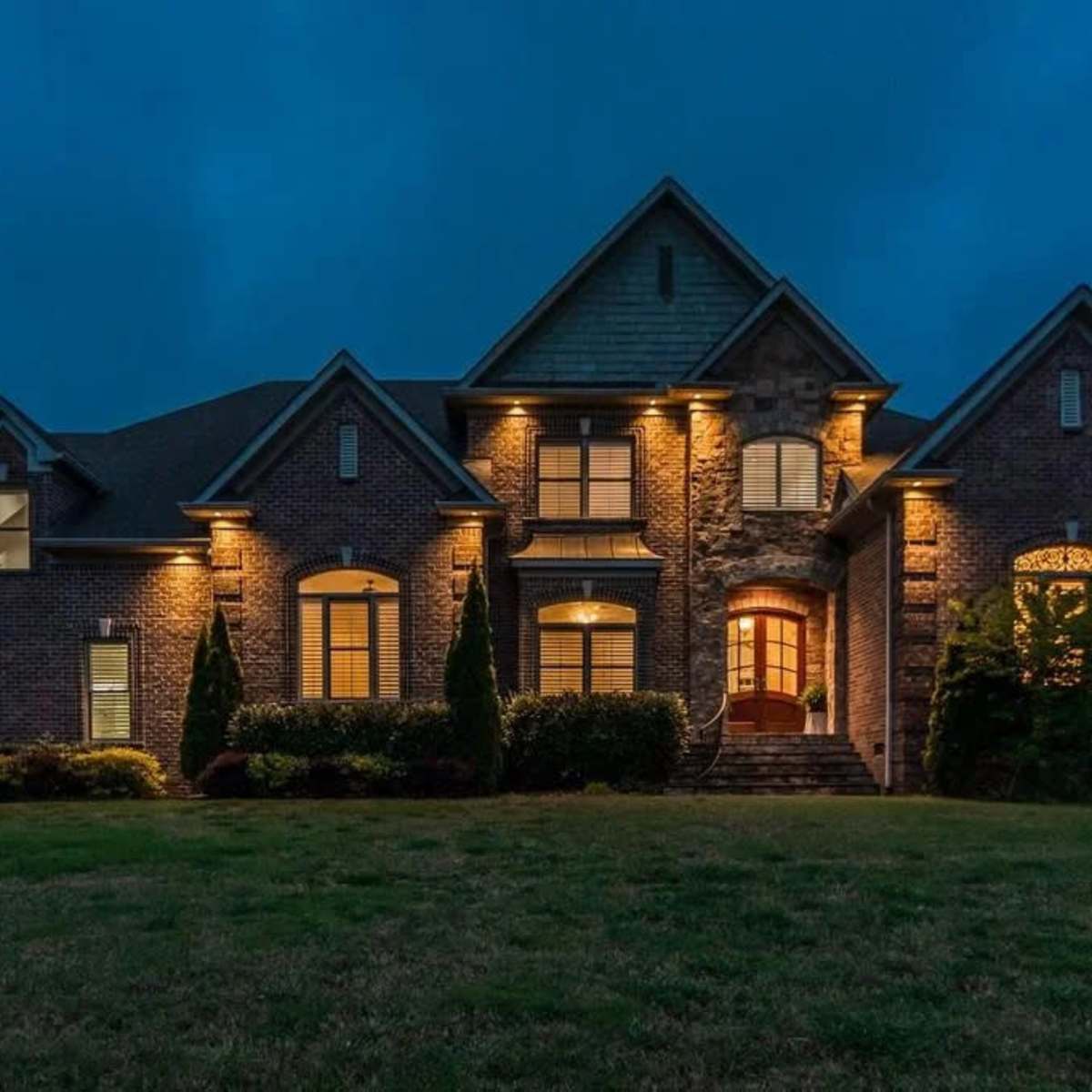 A large brick house with multiple gabled roofs is illuminated by outdoor lights against a twilight sky.