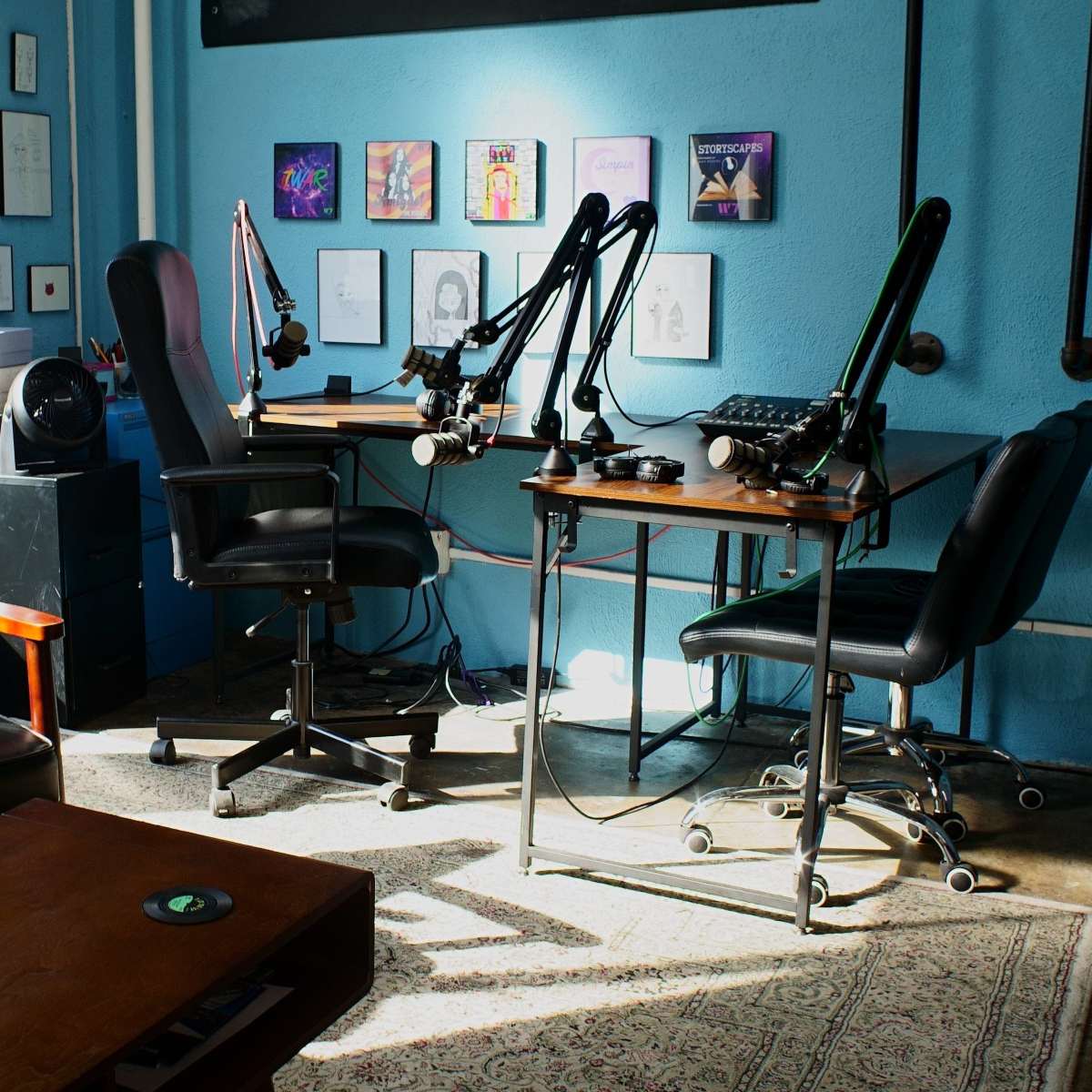 A recording studio with blue walls, two desks equipped with microphones, and a seating area featuring a black chair and wooden coffee table.
