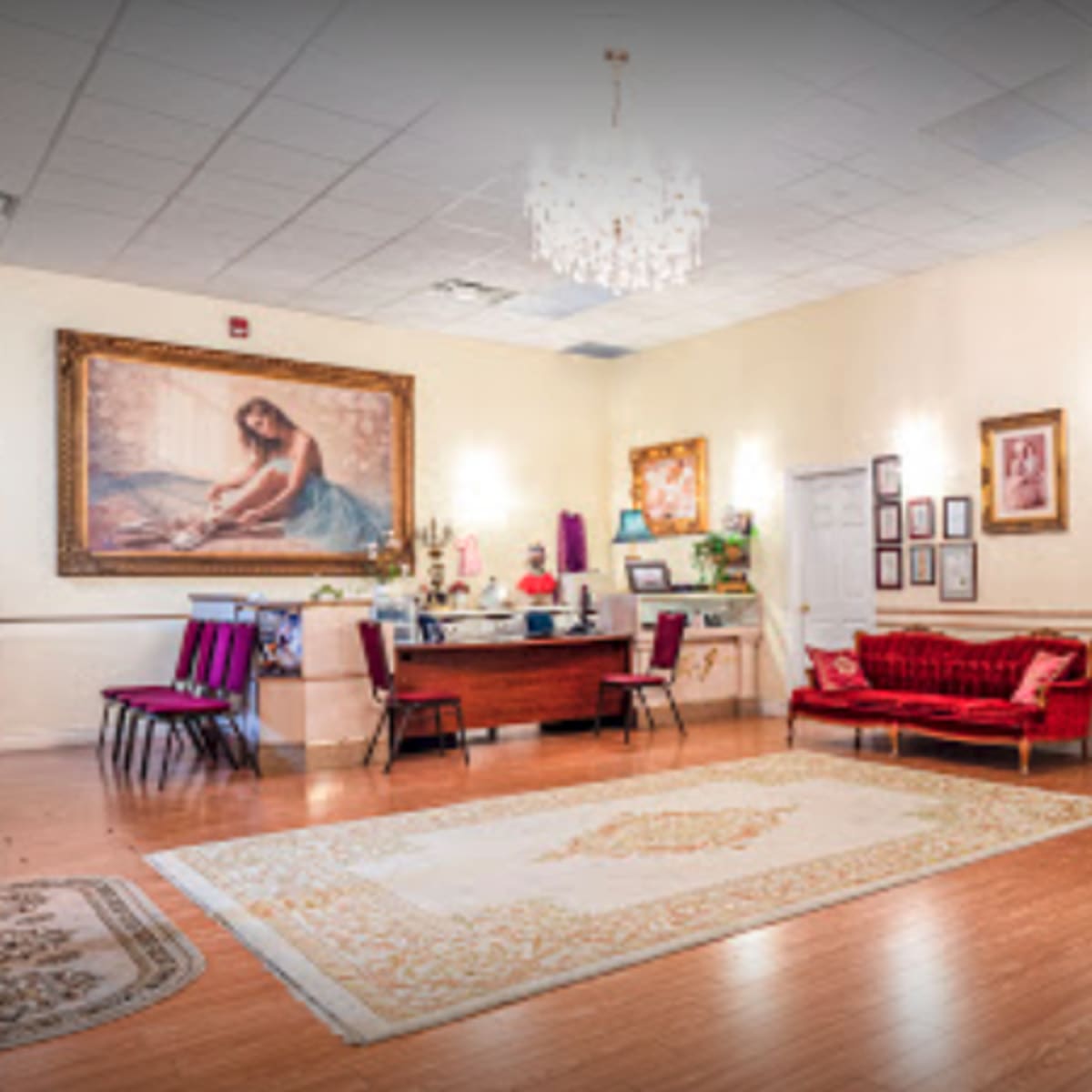 A spacious reception area with wooden flooring, a large painting on the wall, a red velvet couch, and a decorative chandelier.
