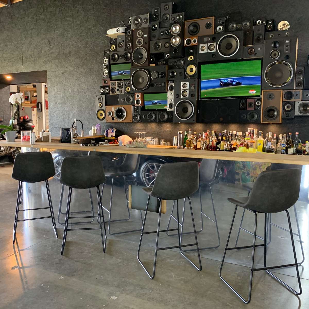 A modern bar area features a long counter with four stools, surrounded by an array of speakers and two televisions mounted on the wall, alongside a collection of liquor bottles.