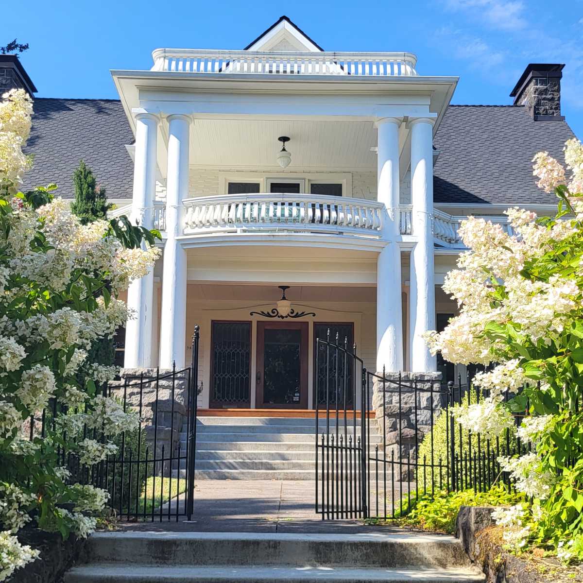 A grand house with white columns stands at the top of a stone staircase, surrounded by blooming hydrangeas and framed by a decorative wrought-iron gate.