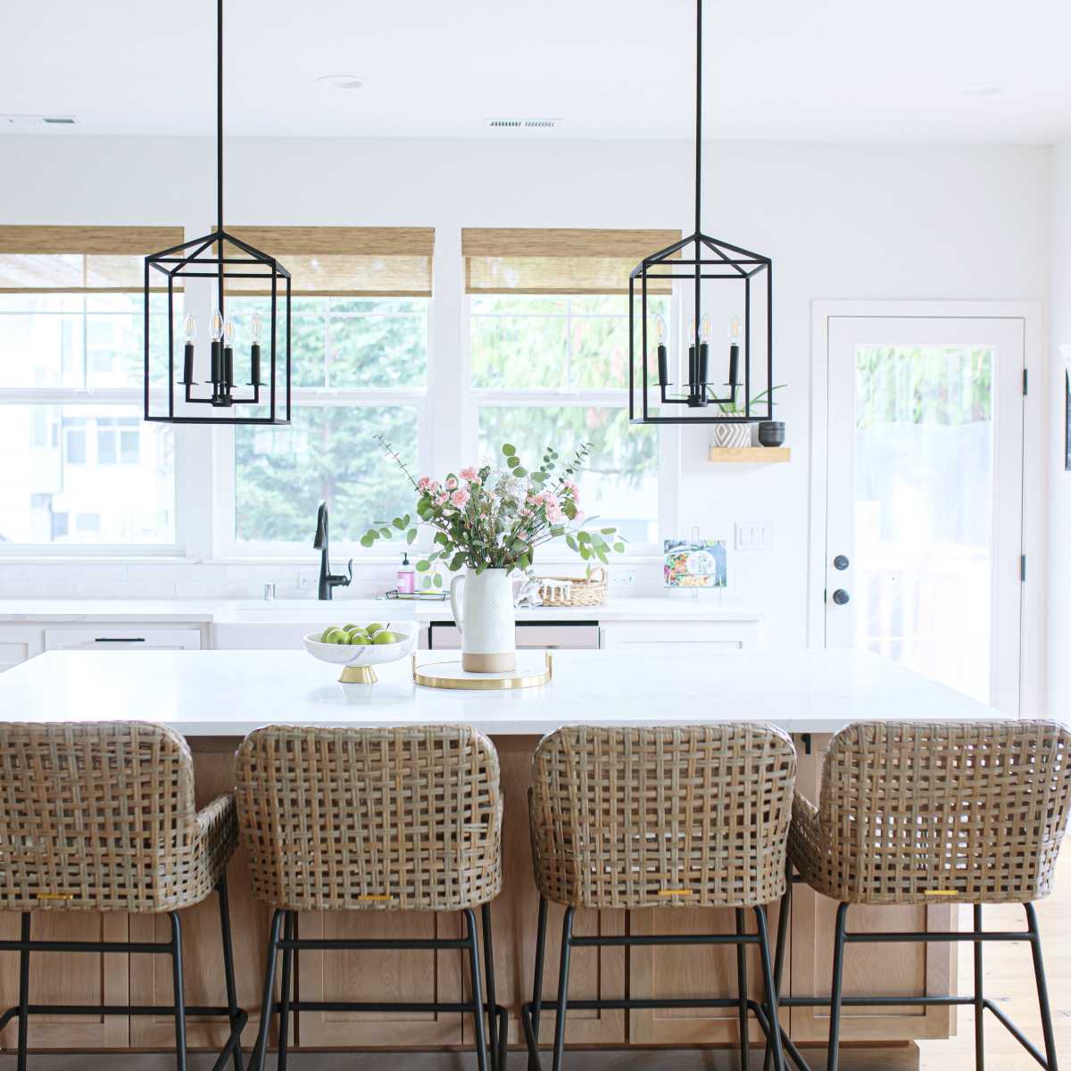 A modern kitchen features a large island with four rattan bar stools, surrounded by windows and pendant lights.