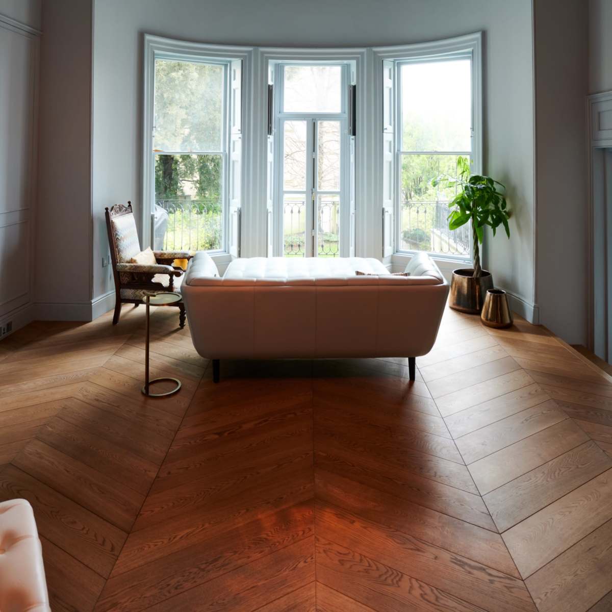 A modern living room features a white sofa, a wooden chair, large windows allowing natural light, and pendant lights above a herringbone-patterned wooden floor.