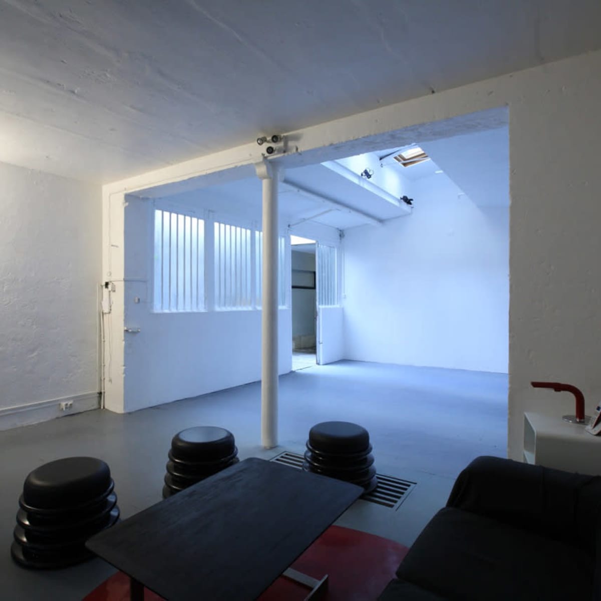 A minimalist, open space with concrete walls, a gray floor, and a few black stools arranged around a low table.