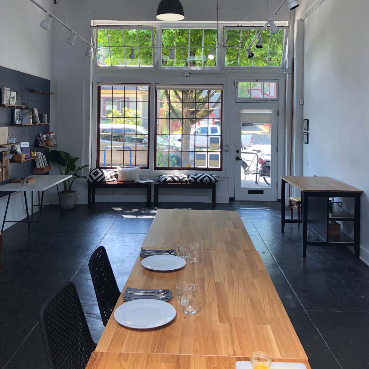 An interior of a modern space featuring a long wooden table with place settings, black chairs, and large windows allowing natural light to enter, showcasing a view of greenery outside.
