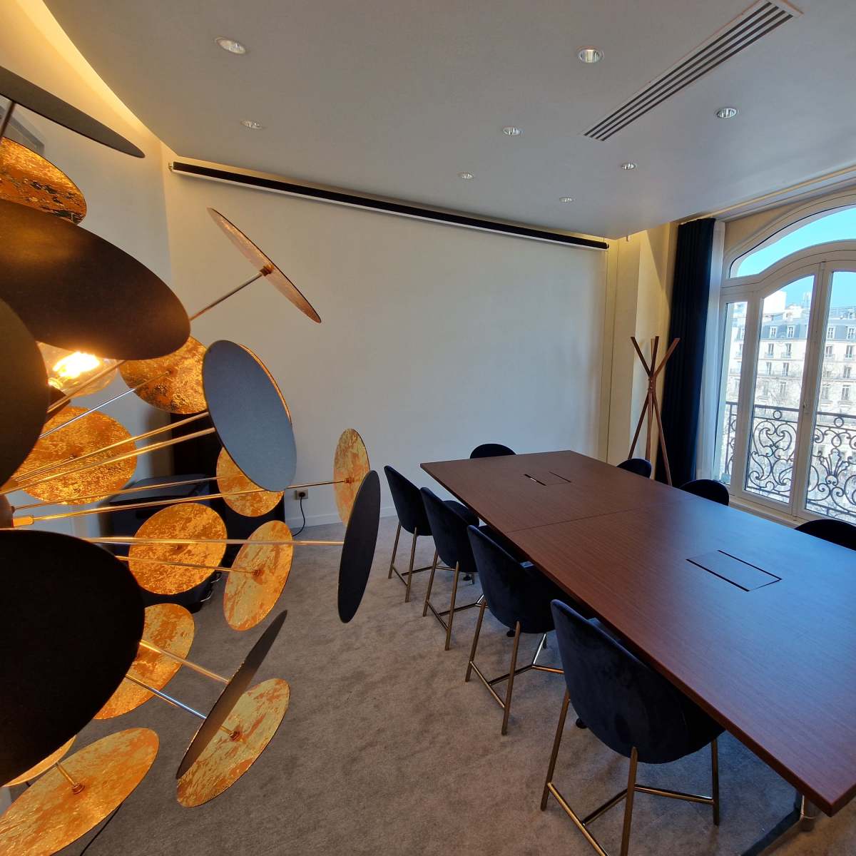 A modern conference room with a large table surrounded by black chairs and a decorative wall feature next to a window overlooking a balcony.